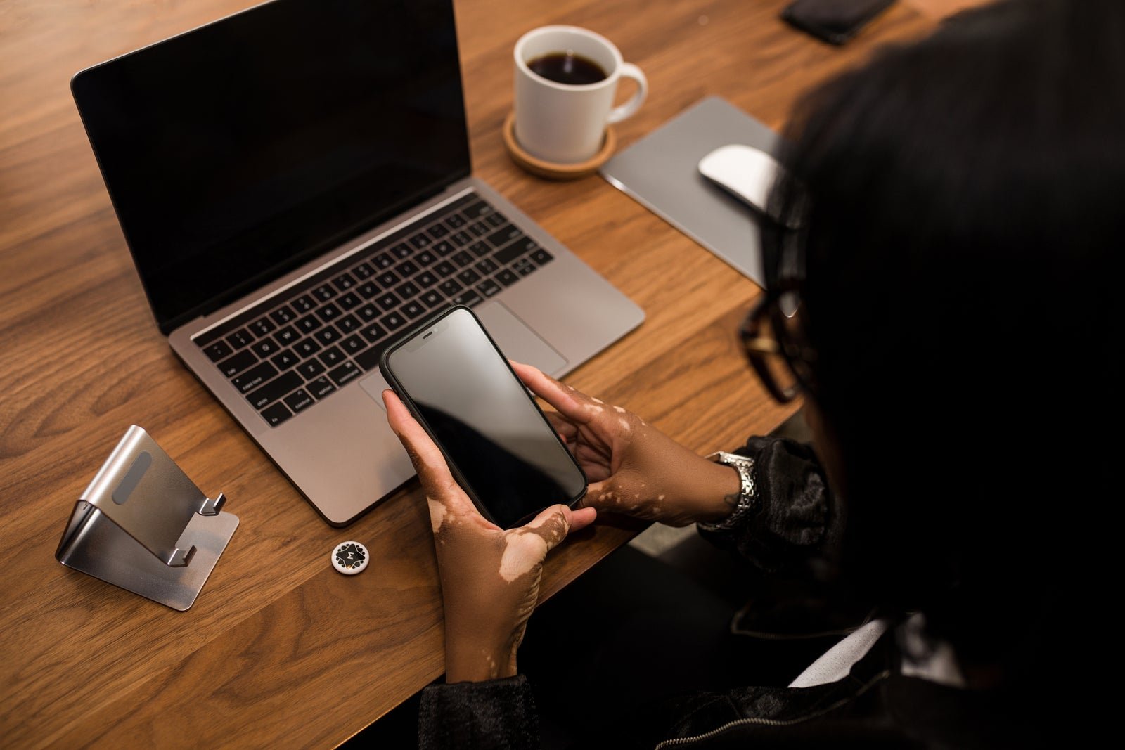 Elderly woman's hand holding a phone over a desk with a laptop, demonstrating technology use at a workspace.
