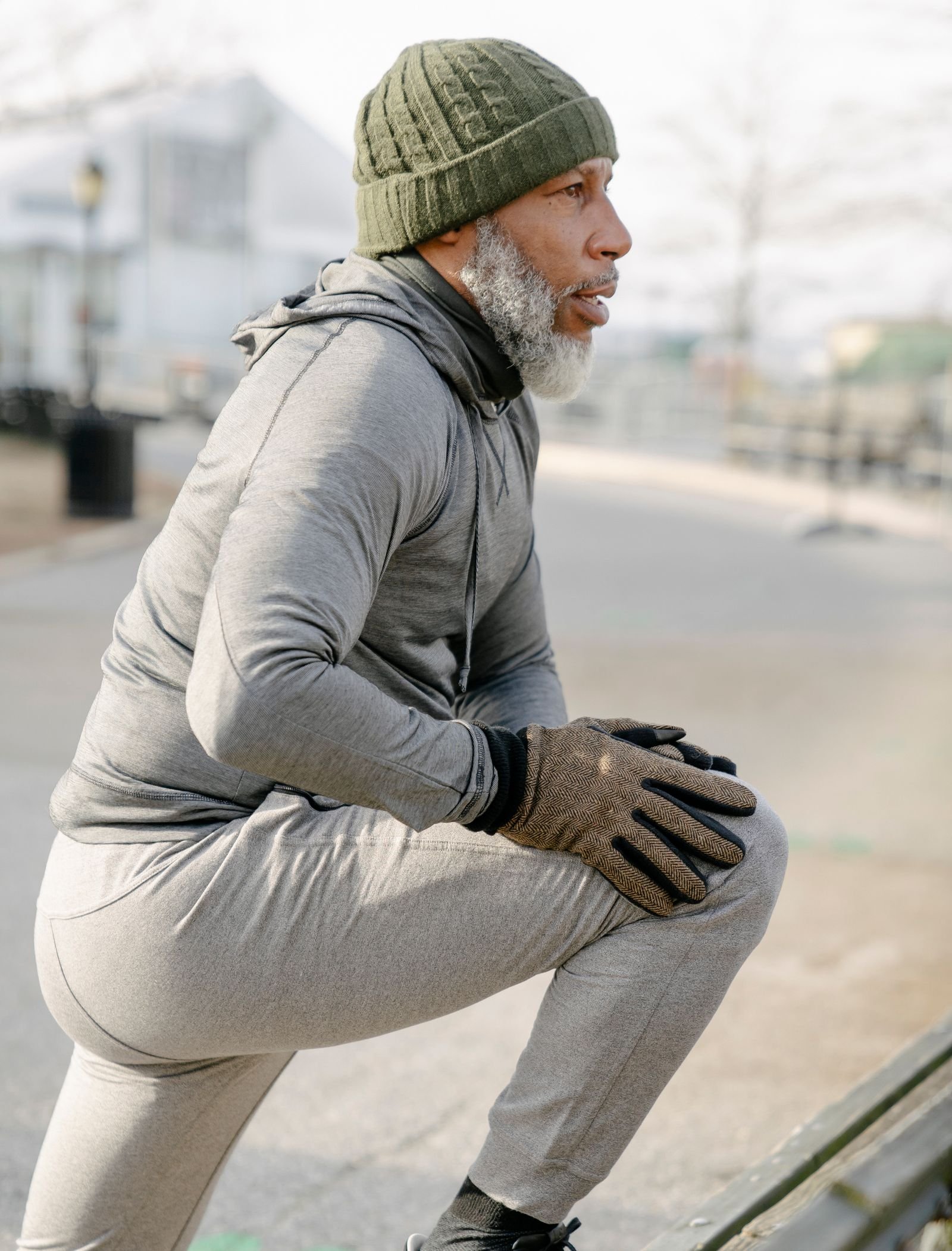 A senior Black man with gray hair and a beard stretches his leg outdoors in an urban park, wearing dark sportswear and a knit hat, warming up before a workout.
