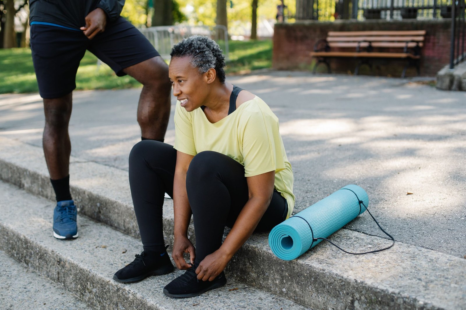Mature Black woman tying her shoes on outdoor steps next to a yoga mat, preparing for exercise