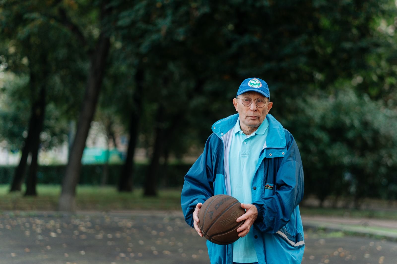 Older Asian man in a blue tracksuit and cap holding a basketball in a park