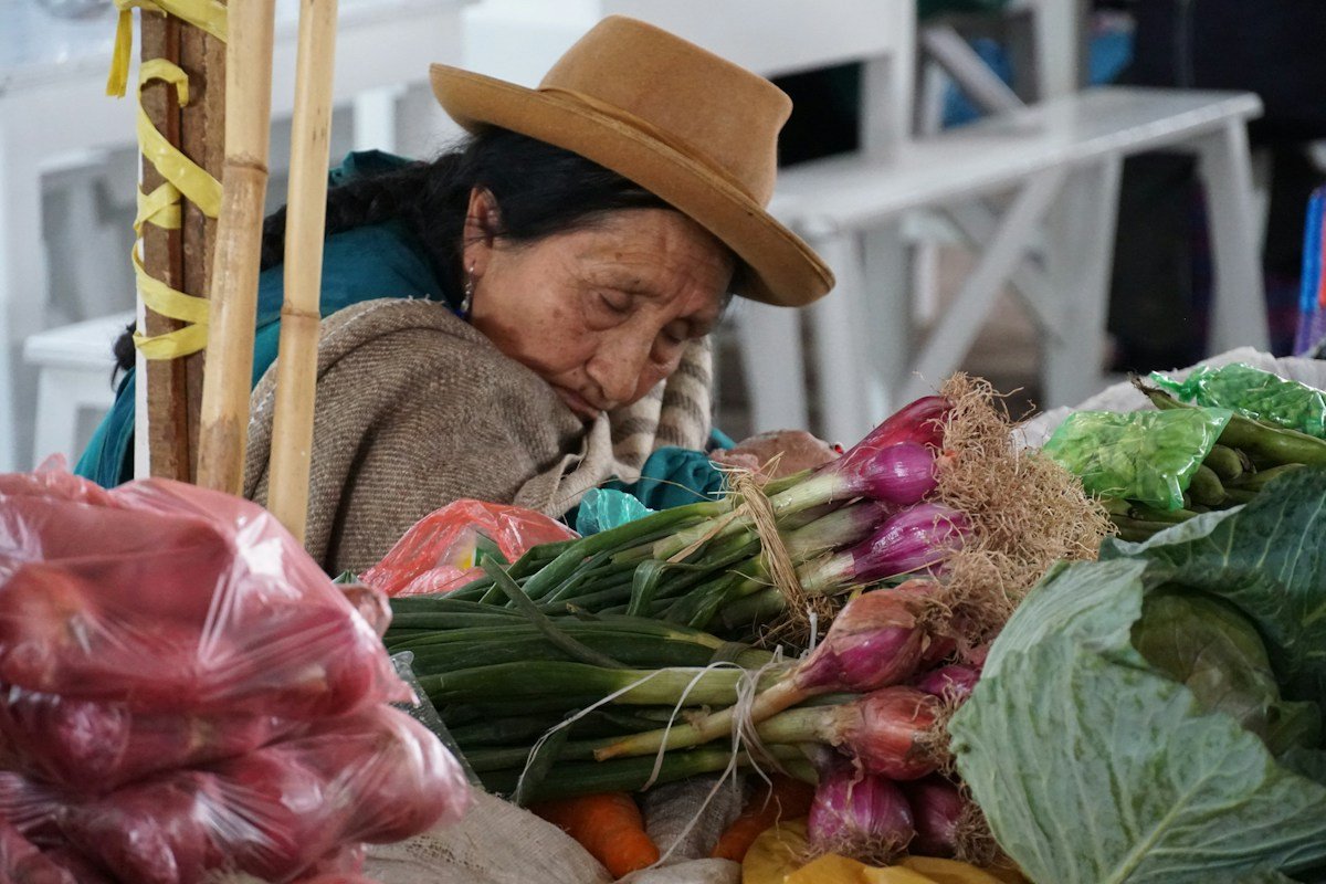 Older woman surrounded by fresh vegetables at an outdoor market, choosing produce for a bone-healthy meal