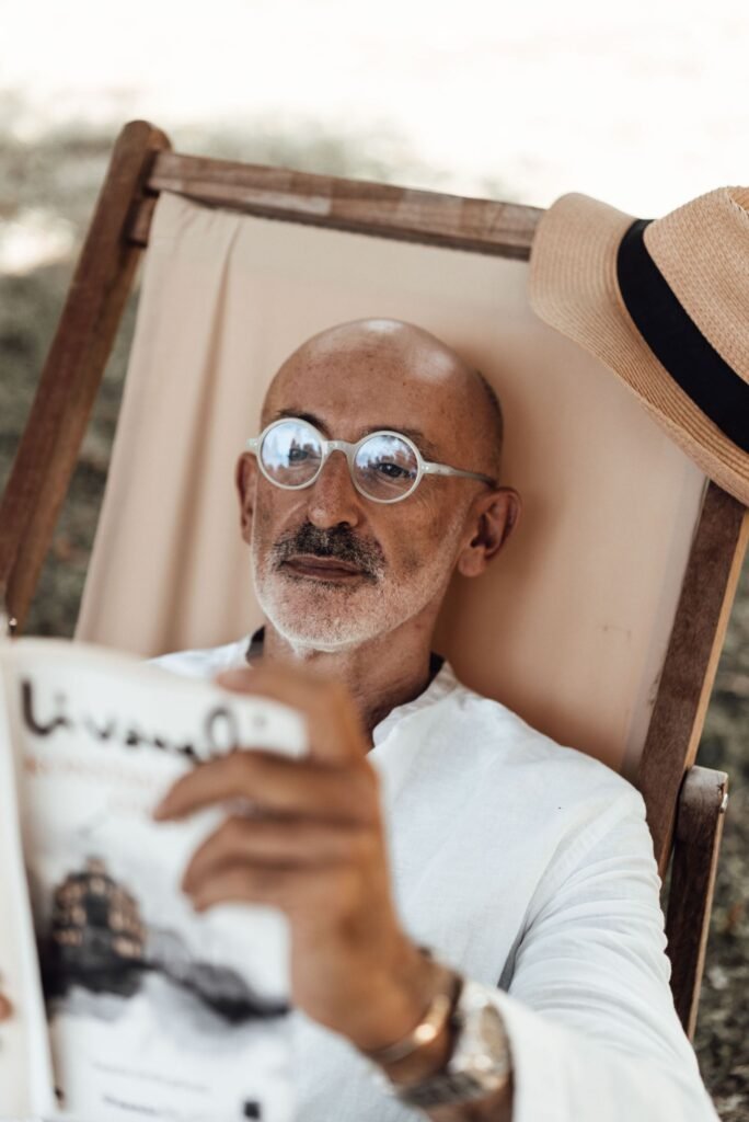 Older man with a gray beard and round glasses reading a magazine outdoors