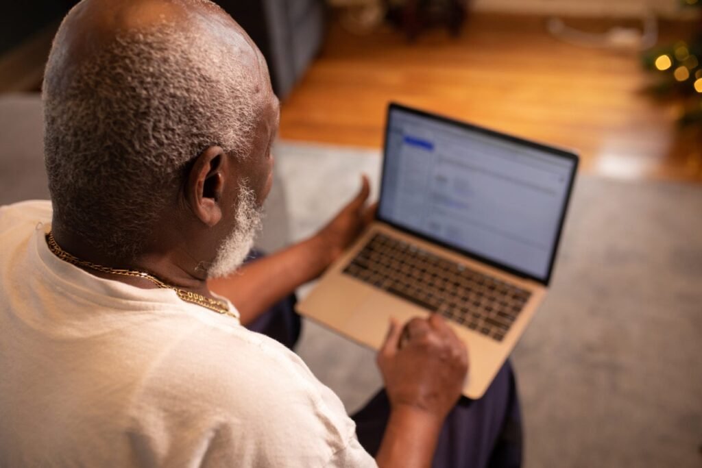 Older man with white hair and beard sitting on a couch with a laptop and phone