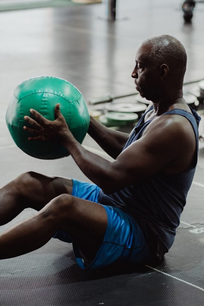 Senior Black man seated on a gym floor gripping a green medicine ball