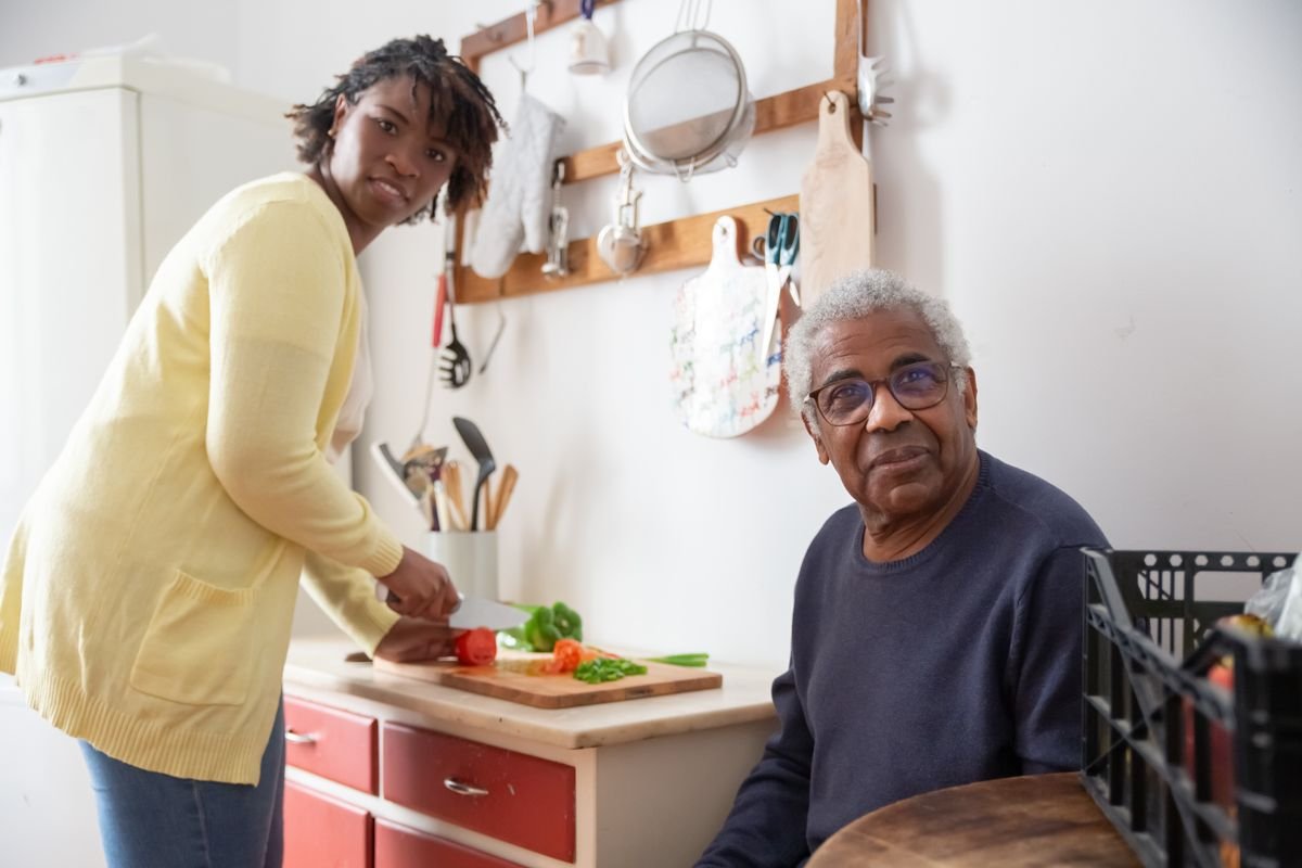 Older Black man sitting in a kitchen while a younger woman prepares fresh vegetables